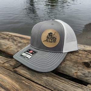 May include: Gray and white trucker hat with a brown leather patch that says "City of St. Clair" with a silhouette of a person fishing.