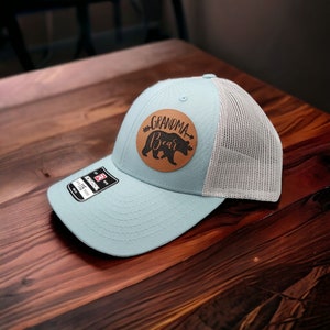 May include: A light blue trucker hat with a brown leather patch that says "Grandma Bear" with a black bear silhouette inside the patch.