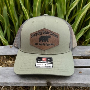 May include: A green and brown camouflage trucker hat with a brown leather patch that says "Sleeping Bear Dunes Michigan" with a black bear silhouette. The hat has a black mesh back and a Richardson 112 adjustable strap.