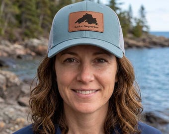 May include: A light blue baseball cap with a mesh back and a rectangular brown leather patch on the front. The patch features a black silhouette of Lake Superior and the text Lake Superior printed in a clear, dark font.