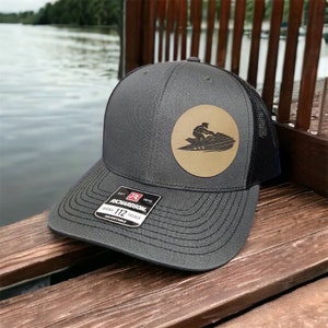May include: A gray and black trucker hat with a brown leather patch featuring a silhouette of a person riding a jet ski. The patch is circular and has a white outline. The hat has a black mesh back and a gray front with a Richardson 112 label.