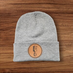 May include: Gray beanie with a brown leather patch featuring a stylized illustration of a piston with a face.