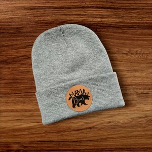 May include: Grey beanie with a brown leather patch that says "Mama Bear" with a black silhouette of a bear inside the patch.