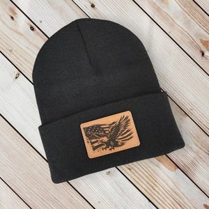 May include: Black beanie hat with a rectangular leather patch. The patch displays an eagle with outstretched wings and the American flag. The hat is made of a soft, knitted material.