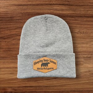 May include: Gray knit beanie with a brown leather patch that says "Sleeping Bear Dunes Michigan" and has a silhouette of a bear.