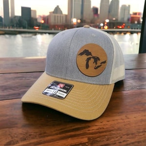 May include: A gray and tan trucker hat with a brown leather patch featuring an outline of the Great Lakes. The hat has a mesh back and the words "Richardson 112 Trucker" on the front.