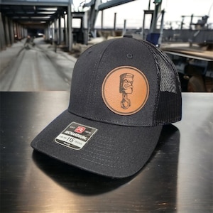 May include: Black trucker hat with a brown leather patch featuring a cartoon piston with a face. The patch is round and has a white outline. The hat is a Richardson 115 Trucker.