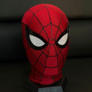 May include: A red and black Spider-Man mask with white mesh eye coverings. The mask features a webbed pattern and is displayed on a black stand. The mask is a replica of the superhero's iconic headwear.