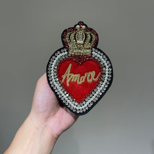 May include: A red felt heart with a gold crown and pearl embellishments. The heart has the word "Amore" embroidered in gold thread.