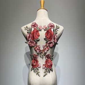 Può includere: Un paio di applicazioni floreali ricamate di colore rosso e verde con un motivo di rosa dettagliato. Le applicazioni sono fissate su uno sfondo di tessuto bianco.