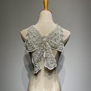 May include: A decorative white bow-shaped embellishment, densely covered in small, round, pearl-like beads. The bow is attached to a sheer, light-colored fabric, creating a layered effect. The design is displayed on a mannequin, showcasing its shape and texture.