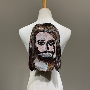 Puede incluir: Un parche de planchado de lentejuelas marrón y negro que representa la cara de un hombre con el pelo largo y barba.