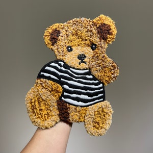 以下が含まれることがあります： 黒と白のストライプのシャツを着た茶色と黄色のテディベアのアプリーケ。クマは悲しい表情をしていて、ふわふわの生地でできています。