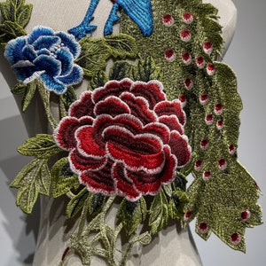 May include: Embroidered floral applique featuring a large red peony, a blue flower, and peacock feather details. The design includes green leaves and stems, with intricate stitching and a textured appearance. The applique is likely for clothing or craft projects.