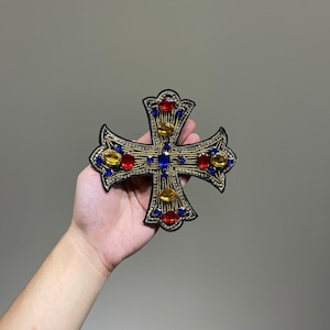 Może przedstawiać: Czarny krzyż z pozłacanymi krawędziami i kolorowymi kryształami. Krzyż jest ozdobiony niebieskimi, czerwonymi i żółtymi kryształami.