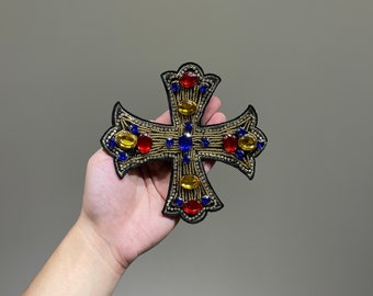 May include: A black cross with gold trim and colorful rhinestones. The cross is decorated with blue, red, and yellow rhinestones.