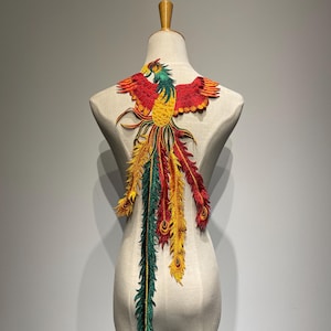 Puede incluir: Aplique bordado de fénix rojo, amarillo y verde con largas plumas de cola que fluyen. El aplique está unido a un fondo de tela blanco.
