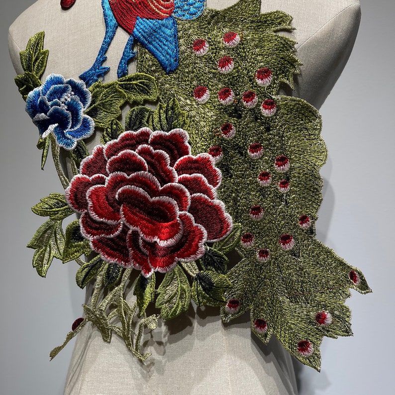 May include: Embroidered floral applique featuring a peacock and peony design. The peacock has blue and red details, while the peony is a deep red with white accents. Green leaves and a blue flower complete the design.
