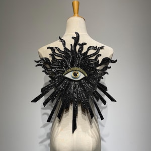 Puede incluir: Una pieza decorativa negra en forma de sol con un ojo en el centro, exhibida en un maniquí. El ojo tiene un iris azul y detalles dorados. El diseño de sol presenta rayos puntiagudos y elementos curvos.