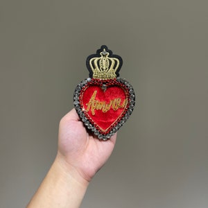 Könnte beinhalten: Ein rotes Samtherz mit einer goldenen Krone und Strasssteinen, mit dem Wort "Amore" in Gold gestickt.