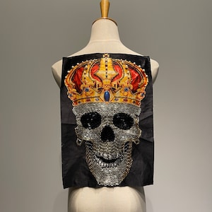 Peut inclure: Un patch en tissu noir avec un crâne argenté portant une couronne dorée avec des accents rouges et bleus. Le crâne est orné de strass.