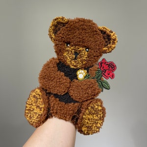 Op de afbeelding: Een bruine teddybeer die een rode roos met groene bladeren vasthoudt. De beer heeft een zwarte neus en een pootprint op zijn borst.