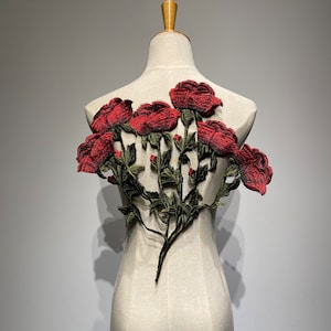 以下が含まれることがあります： マネキンに飾られた、緑の葉と茎が付いた赤いバラのアップリケ。 花柄は、複数の大きく質感のあるバラの花を特徴としています。 アップリケは、衣類やクラフトプロジェクト用である可能性があります。