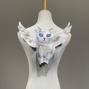 May include: A white cat with blue eyes and wings, embroidered on a light brown fabric.