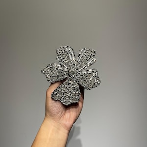 Könnte beinhalten: Eine silberne Brosche in Blumenform mit klaren Strasssteinen. Die Brosche ist aus Metall und hat ein filigranes Design.