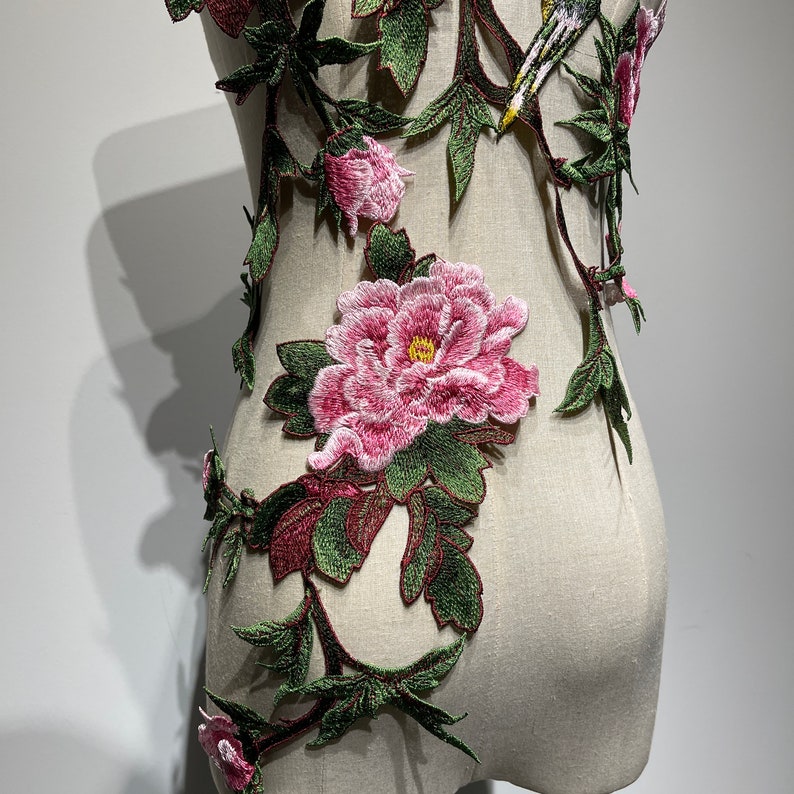 Pode incluir: Uma aplica&ccedil;&atilde;o floral delicada com pe&ocirc;nias cor-de-rosa e folhas verdes. A aplica&ccedil;&atilde;o &eacute; bordada em um tecido branco e seria perfeita para adicionar um toque de eleg&acirc;ncia a uma roupa ou projeto de decora&ccedil;&atilde;o de casa.