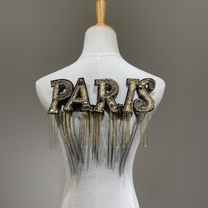 Pode incluir: Uma aplicação "PARIS" com contas douradas e pretas com franjas de corrente dourada.