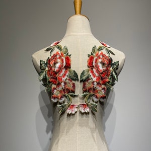 May include: Embroidered floral applique featuring large red and white peony-like flowers with green leaves. The applique is designed to be sewn onto fabric, with a detailed and textured appearance, perfect for embellishing garments.