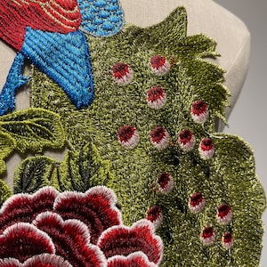 May include: Embroidered applique featuring a bird and floral design. The bird has red and blue feathers, while the flowers are a deep red with white edges. The leaves are green with black accents, creating a detailed and colorful textile embellishment.