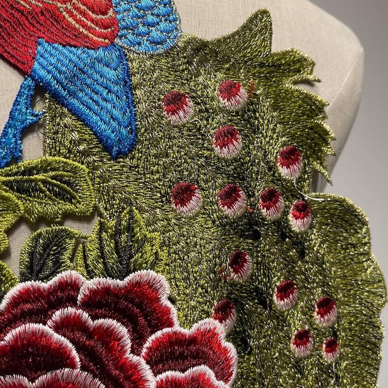 May include: Embroidered applique featuring a bird and floral design. The bird has red and blue feathers, while the flowers are a deep red with white edges. The leaves are green with black accents, creating a detailed and colorful textile embellishment.