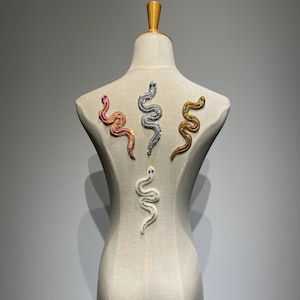 Peut inclure: Un mannequin blanc avec quatre décorations en forme de serpent. Les serpents sont faits de strass et sont de différentes couleurs : rose, argent, or et blanc.
