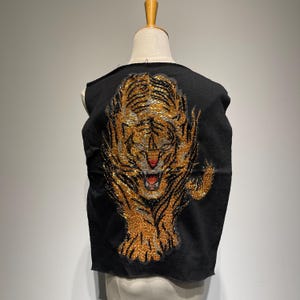 Può includere: Un gilet nero senza maniche con un grande disegno di tigre ricamato sul retro. La tigre è realizzata con perline dorate e nere e ha un'espressione feroce.