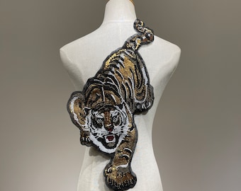 May include: A large, detailed tiger applique with gold and black sequins. The tiger is facing forward with its mouth open and its eyes wide. The applique is perfect for adding a touch of wildness to any project.