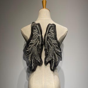 May include: Two black, sheer fabric shoulder pieces, each adorned with a detailed, silver beaded feather design. The embellishments are intricate, with a mix of beads and sequins. The pieces are displayed on a mannequin.