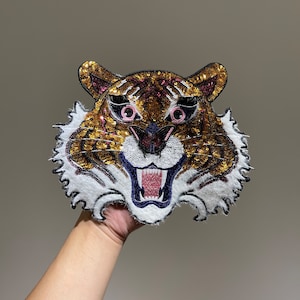 Könnte beinhalten: Ein gold- und braunes Tigergesichts-Applik mit rosa und blauen Details. Der Tiger hat einen weißen Pelzbesatz und ist aus Pailletten gefertigt.