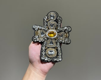 May include: Embroidered cross applique with a black base, adorned with clear and yellow gemstones, and gold-colored beads. The cross features a central yellow stone and is embellished with various shapes of sparkling crystals.