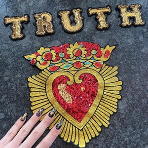 K&ouml;nnte beinhalten: Ein golden besticktes Herz mit einem roten Pailletten-Zentrum und einer Krone oben. Das Herz ist von goldenen Strahlen umgeben. Das Wort "TRUTH" ist in goldenen Buchstaben &uuml;ber dem Herzen geschrieben.