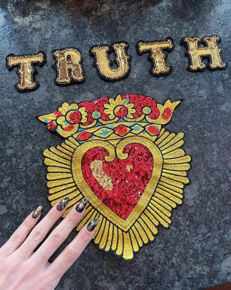 K&ouml;nnte beinhalten: Ein golden besticktes Herz mit einem roten Pailletten-Zentrum und einer Krone oben. Das Herz ist von goldenen Strahlen umgeben. Das Wort "TRUTH" ist in goldenen Buchstaben &uuml;ber dem Herzen geschrieben.