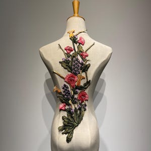Pode incluir: Aplique floral bordado com um arranjo vertical de flores vermelhas, rosa, roxas e amarelas com folhas verdes. O aplique é exibido em um manequim bege, mostrando os detalhes intrincados do design.