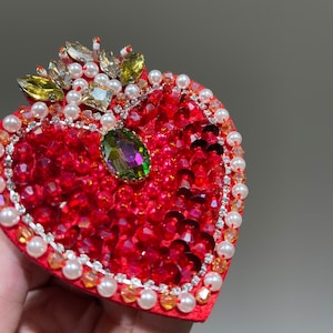K&ouml;nnte beinhalten: Eine rote herzf&ouml;rmige Brosche mit einer Krone aus goldenen und gr&uuml;nen Strasssteinen, umgeben von wei&szlig;en Perlen. Das Herz ist mit roten Pailletten und Strasssteinen bedeckt.