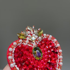 K&ouml;nnte beinhalten: Eine rote herzf&ouml;rmige Brosche mit einer Krone aus Perlen und Strasssteinen. In der Mitte des Herzens befindet sich ein gro&szlig;er gr&uuml;ner und violetter Edelstein.