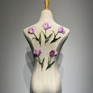 Könnte beinhalten: Eine beigefarbene Schneiderpuppe, verziert mit gestickten lila Tulpen-Applikationen. Die Tulpen haben grüne Stiele und Blätter, die ein Blumenmuster bilden. Die Schneiderpuppe steht vor einem neutralen grauen Hintergrund.