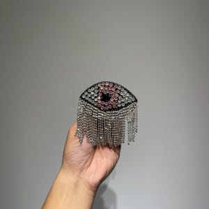 May include: A black eye-shaped brooch with pink and clear rhinestones and a fringe of silver chains.