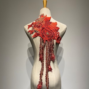 Könnte beinhalten: Rote gestickte Applikation mit floralem Muster und goldenen Akzenten. Die Applikation zeigt eine große Blume mit einer kleineren Blume und Blättern. Die Applikation ist auf einem weißen Stoffgrund befestigt.