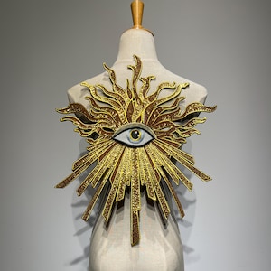 May include: A gold and brown sunburst-shaped applique with an eye in the center, displayed on a mannequin. The eye is blue, gray, and white. The sunburst has detailed rays and a textured appearance.