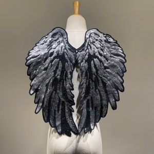 Könnte beinhalten: Ein Paar schwarze und silberne Pailletten-Engel Flügel. Die Flügel sind an einem Mannequin befestigt und werden vor einem weißen Hintergrund ausgestellt.